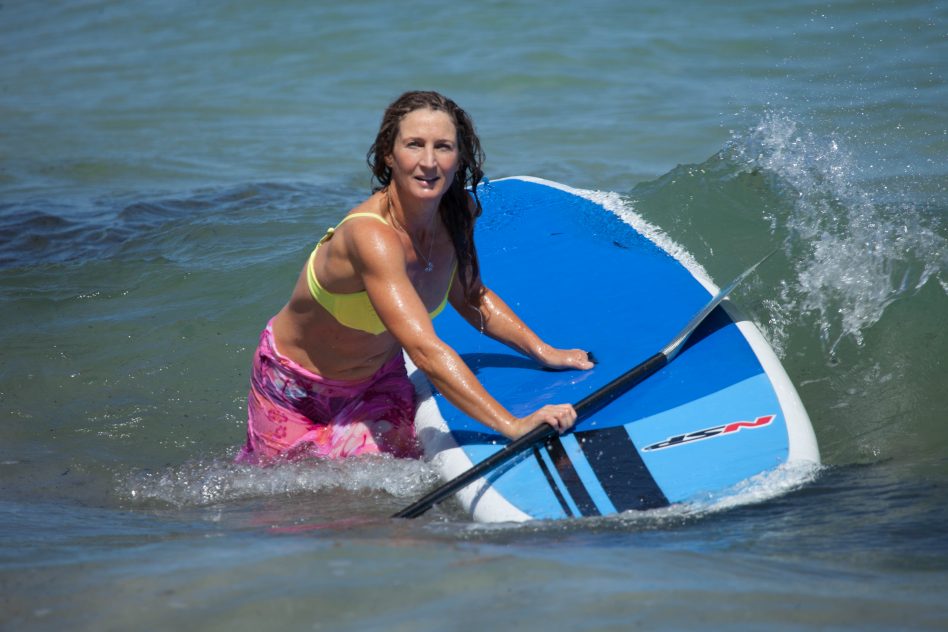 A picture of Maree in the surf standing beside her Paddleboard while a wave is starting to crash behind her