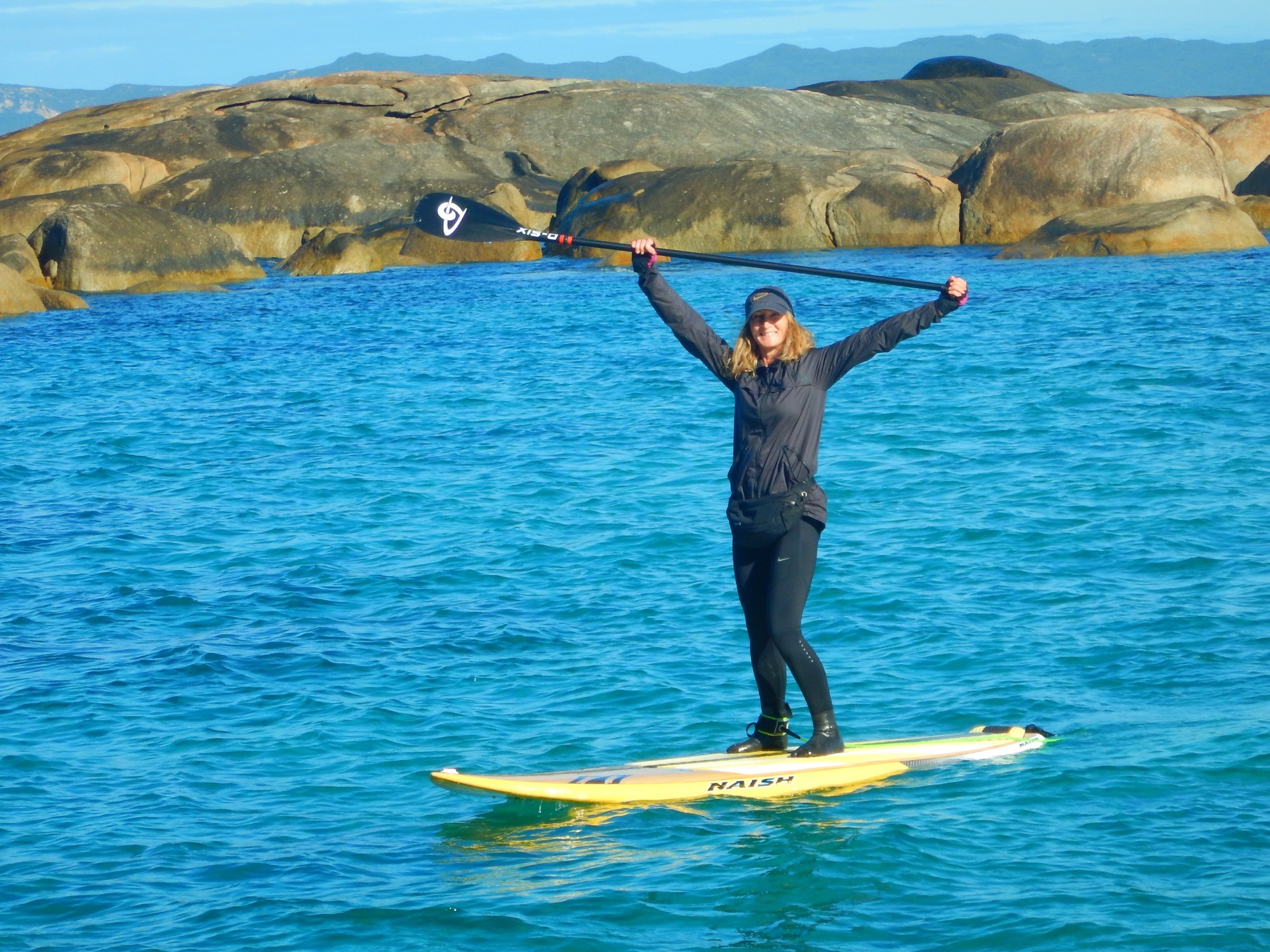 Maree standing on her yellow SUP board with a SUP paddle held high in her hands