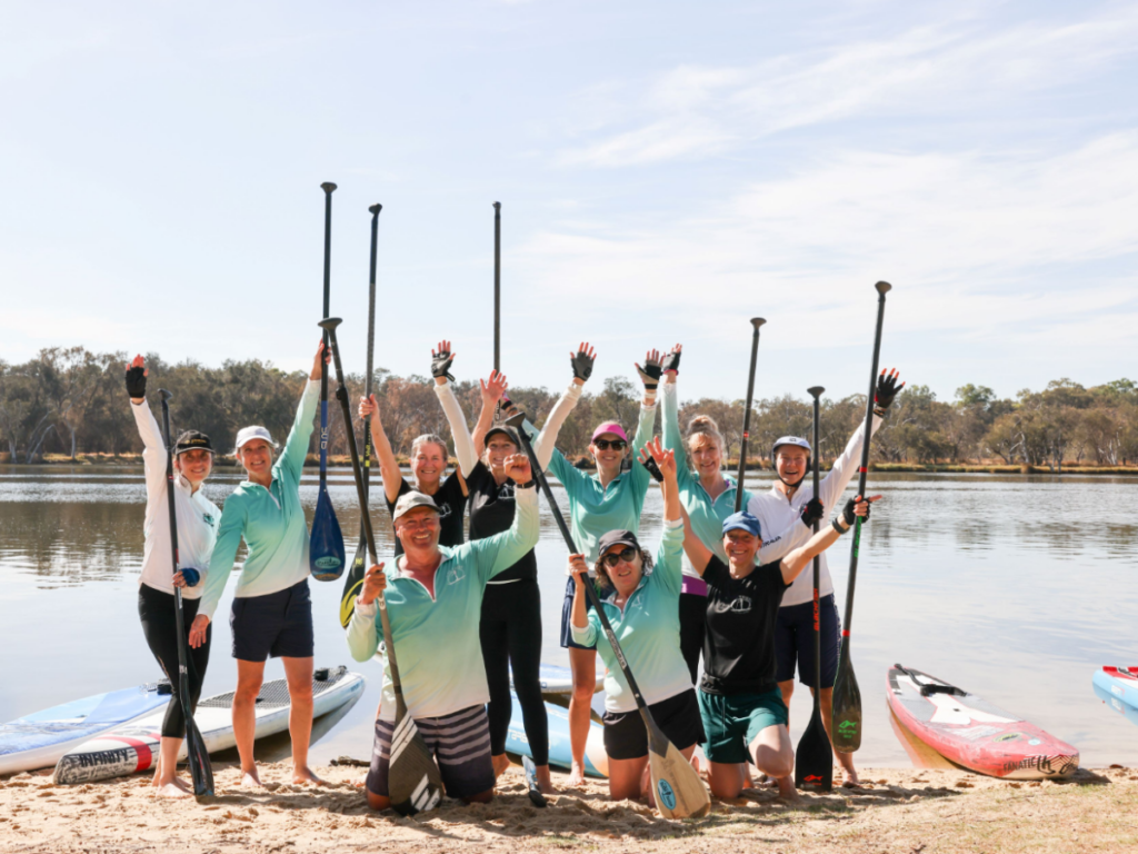 A group of people kneeling on the banks of the swan river with thier paddles up in the air, all wearing SUP TONIC merchandise and looking very happy